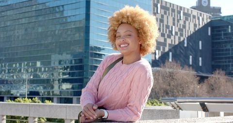 Confident Young Woman Smiling on City Terrace Wearing Pink Sweater with Glass Architecture