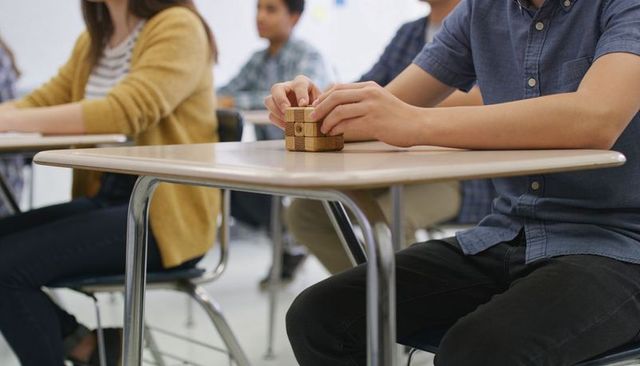 Male student solving wooden cube puzzle at classroom desk while classmates concentrate