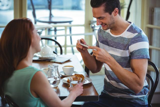 Man taking photo of croissant with mobile phone in café. Ideal for use in lifestyle blogs, social media content, food photography tutorials, and advertisements for cafés or restaurants.