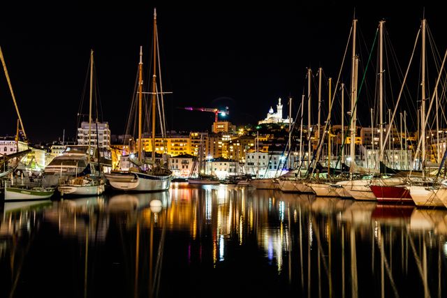 Capturing a vibrant night scene of a marina with numerous yachts docked, reflecting off the calm water below. The well-lit buildings and a historical monument in the background enhance the charm of the coastal city. Suitable for use in travel brochures, tourism websites, urban planning presentations, and lifestyle magazines promoting night life by the sea.
