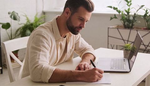 Bearded man writing at sunlit minimalist home workspace with laptop and plants