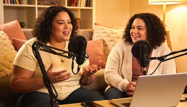 Hispanic podcast duo recording cozy living room conversation with microphones and laptop