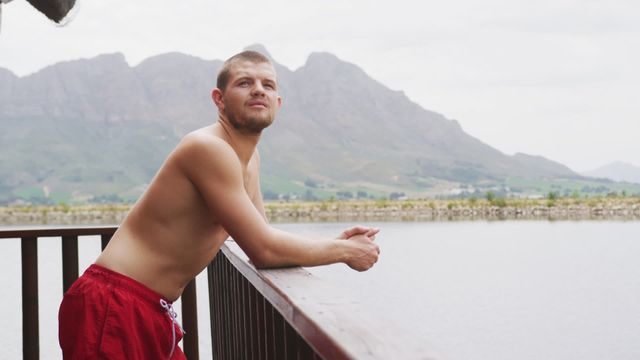 A man is standing on a cabin balcony, wearing red swim trunks and enjoying the view of a mountain river. Perfect for themes related to travel, relaxation, outdoor adventures, leisure activities, and peaceful retreats. Useful for print and digital content in tourism advertisements or lifestyle blogs emphasizing nature and escape.