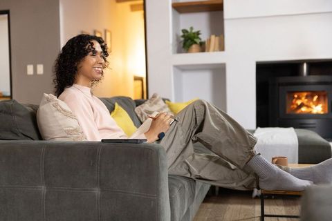 Non-binary person journaling on gray sofa by fireplace, cozy relaxed home lifestyle