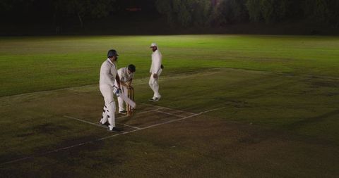 Floodlit night cricket players preparing batting crease and inspecting stumps on pitch