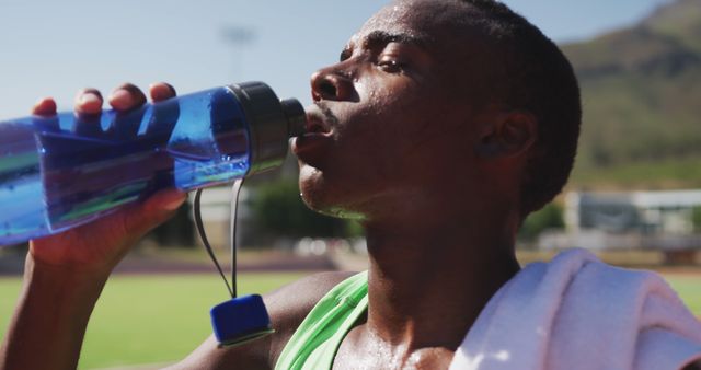 Tired african american male athlete drinking water at empty stadium. Sport, atletics and fitness, unaltered.