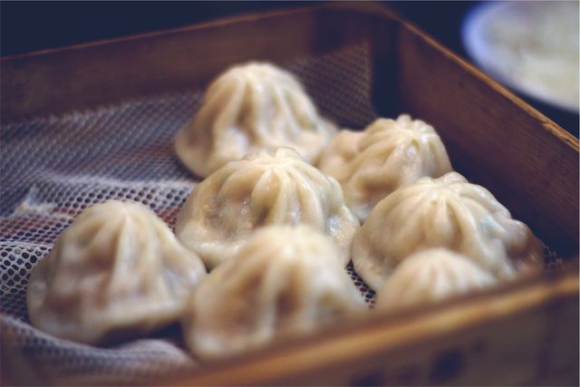 Close-up of steamed dumplings served in a traditional wooden bamboo basket, typically found at Chinese dim sum. Exhibits traditional culinary art with intricately folded dough, suggesting freshness and homemade quality. Ideal for use in culinary blogs, food packaging, restaurant menus, and cultural articles focusing on Asian cuisine.