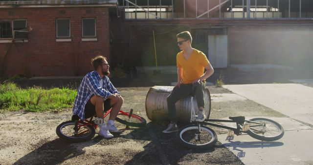 Two young men are sitting on their bicycles in an urban industrial area during a sunny day. This can be used for themes of friendship, urban exploration, youth lifestyle, or outdoor activities. Relevant for promotions in an urban setting or for articles discussing trends among young people.