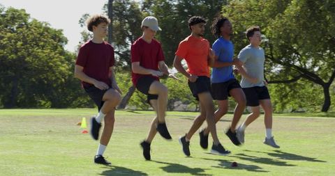 Diverse male team performing high-knee and butt-kick drills on grass for fitness training