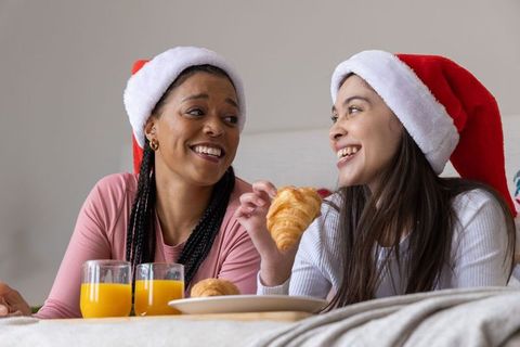 Diverse women wearing santa hats sharing croissants and orange juice on cozy holiday bed