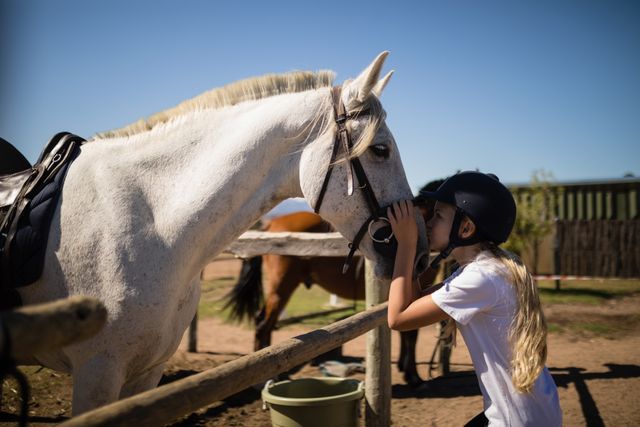 Young girl wearing helmet kissing white horse at ranch on sunny day. Ideal for themes of childhood, animal bonding, equestrian activities, rural life, and outdoor leisure. Suitable for use in advertisements, blogs, educational materials, and social media posts related to horses, pets, and countryside living.