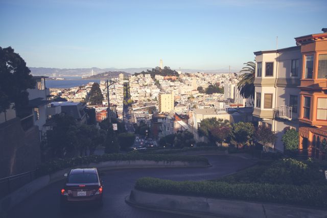 Scenic view of Lombard Street in San Francisco during sunset, showcasing the city's iconic winding road and urban landscape. The golden light captures the architecture of residential buildings, and Coit Tower is visible in the distance. Ideal for promoting tourism in San Francisco or illustrating urban scenery and travel destinations.