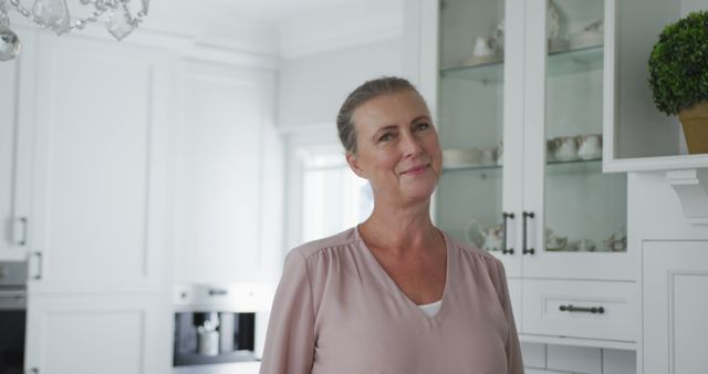 Mature woman standing in a bright, modern kitchen with white cabinets and natural lighting, wearing a pink blouse. She is smiling and looking content, demonstrating comfort and happiness at home. This image might be used for lifestyle articles, advertisements promoting home goods, or campaigns focusing on positive, everyday living.