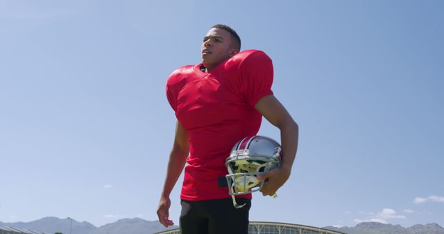 Young American football player wearing red jersey holding helmet under clear sky. Ideal for use in promotional materials for sports teams, motivational posters, fitness and training content, and advertisements for sporting goods.