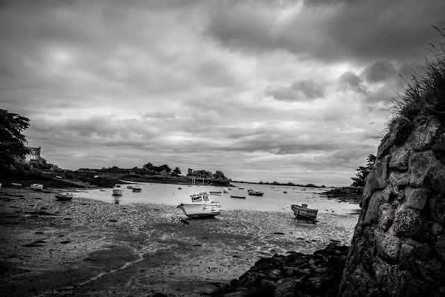 Black-and-white seascape showing multiple small boats stranded on a rocky shoreline during low tide under a cloudy sky. Suitable for concepts of tranquility, dramatic landscapes, and natural coastal scenery. Ideal for use in travel articles, nature-themed blogs, and coastal environmental studies.