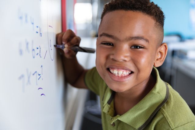 Young African American boy smiling while writing on a whiteboard in a classroom. Ideal for educational materials, school promotions, and content focused on childhood learning and happiness.