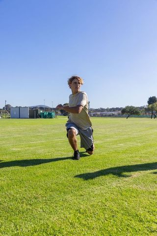 Young male athlete lunging on sunlit grass field under clear blue sky for sports training