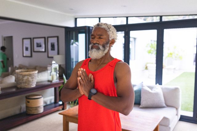 Senior African American man meditating with hands clasped in a peaceful home environment. Ideal for use in articles or advertisements related to fitness, active lifestyle, mental health, mindfulness, and wellness. Can be used to promote yoga classes, meditation apps, or healthy living products.
