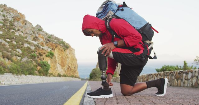 Biracial man kneeling to check prosthetic leg and shoe, trekking with backpack on coastal road. Long distance walking, fitness, challenge, disability, nature and healthy outdoor lifestyle.