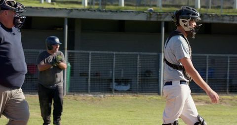 Baseball catcher walking with protective gear, umpire by dugout and teammate clapping