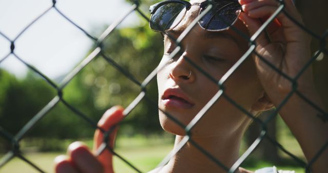 Biracial woman wearing sunglasses and holding chain-link fence at park - Download Free Stock Photos Pikwizard.com