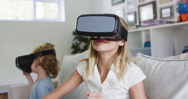 Two children are playing and enjoying virtual reality experiences together in a living room. One child is sitting comfortably on a couch while the other is engaged with the VR headset. The living room has a cozy and modern feel with family photos and shelves in the background. This can be used for educational materials, technology advertisements, gaming promotions, or articles on the impact of virtual reality on children.