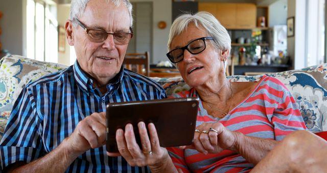 A senior Caucasian couple is engaged with a tablet, sharing a moment of digital interaction in a cozy home setting. Their expressions suggest a mix of concentration and enjoyment as they navigate the device together.