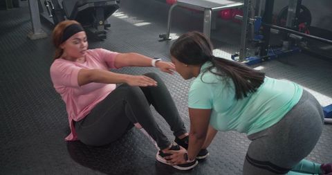 Diverse mid-adult women performing partnered sit-ups in gym trainer assisting