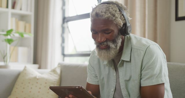 Elderly African American man sitting comfortably on a sofa at home, smiling while listening to music using a tablet and headphones. Suitable for use in articles, advertisements, or promotions related to technology, leisure activities for seniors, modern lifestyle, and home electronics.