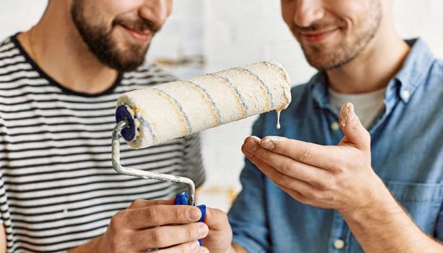Two men smiling holding paint roller dripping paint while renovating interior at home