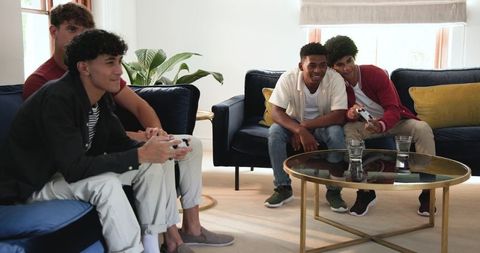 Multiracial young men playing console games on sofa in modern sunlit living room