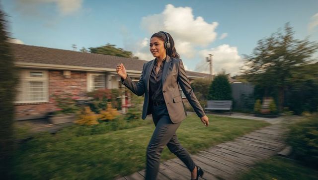 Businesswoman walking through suburban garden wearing over-ear headphones, confident stride