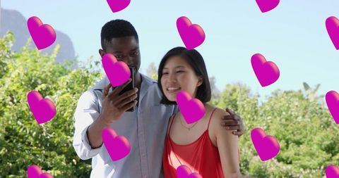 Young couple sharing smartphone surrounded by floating pink hearts in sunny garden