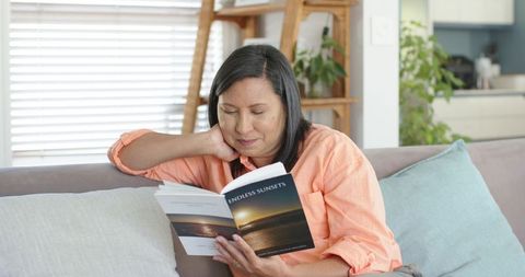 Mature asian woman reading paperback novel on sofa relaxing in modern living room