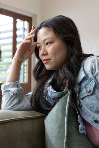 Asian woman sitting on sofa contemplating in denim jacket holding cushion in natural light