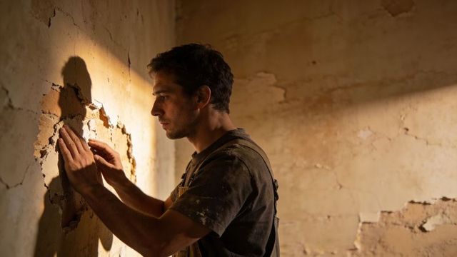 Worker inspecting cracked plaster wall in sunlit room during renovation work and repair