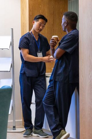 Two male clinicians wearing navy scrubs chatting during coffee break in staff lounge