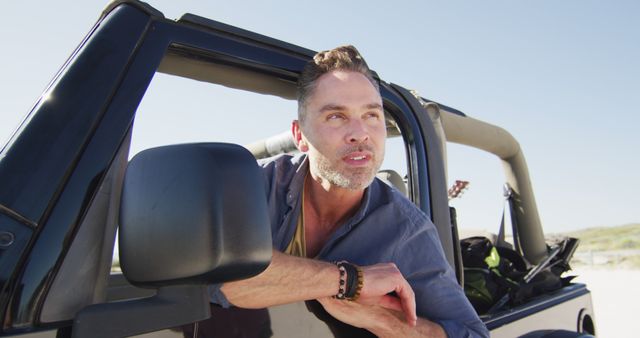 Man leaning out of window of a convertible off-road vehicle, basking in the sunlight. Great for use in travel and adventure promotions, tourism advertisements, recreational activities, and lifestyle blogs. Depicts themes of exploration, freedom, and outdoor adventures.