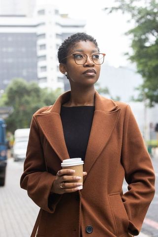 Standing african professional holding coffee, wearing rust coat and tortoiseshell glasses