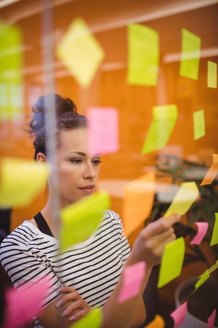 Businesswoman reading colorful sticky notes on glass wall in modern office. Ideal for illustrating concepts of brainstorming, planning, strategy, and productivity in a corporate environment. Useful for business presentations, team collaboration visuals, and project management resources.