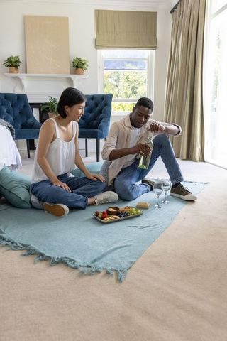 Couple sitting on blanket in living room opening white wine and sharing charcuterie platter