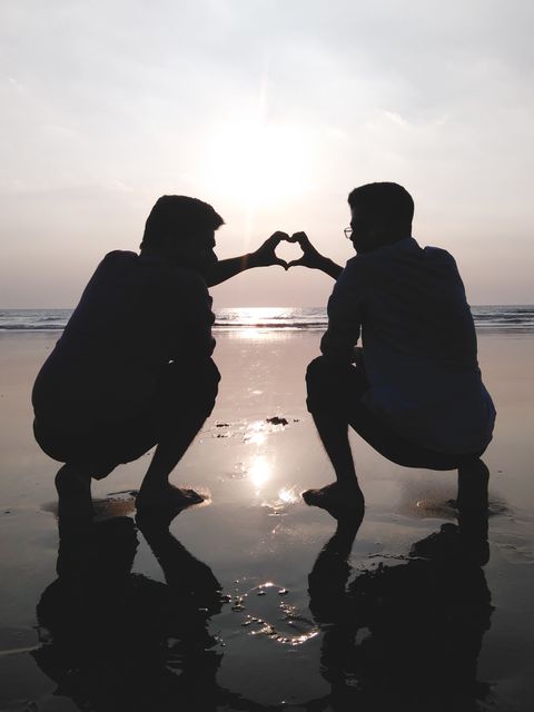 Two people show affection while creating a heart shape with their hands against a sunset background on the beach. Perfect for themes of love, relationships, friendship, and tranquility. Suitable for Valentine's Day, tourism ads, or inspirational content about togetherness and peace.