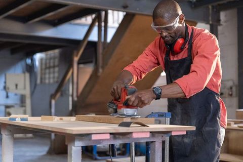 African american craftsman sanding wooden board in workshop wearing apron and safety goggles