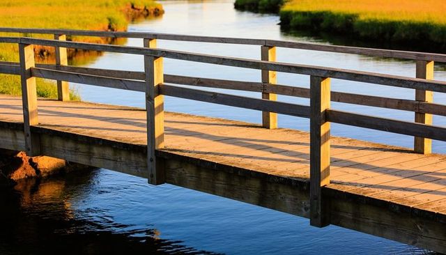 Sunlit weathered wooden footbridge over tranquil marsh creek with reflections