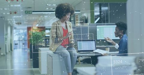 Two coworkers collaborating over data on laptops in open office with digital interface