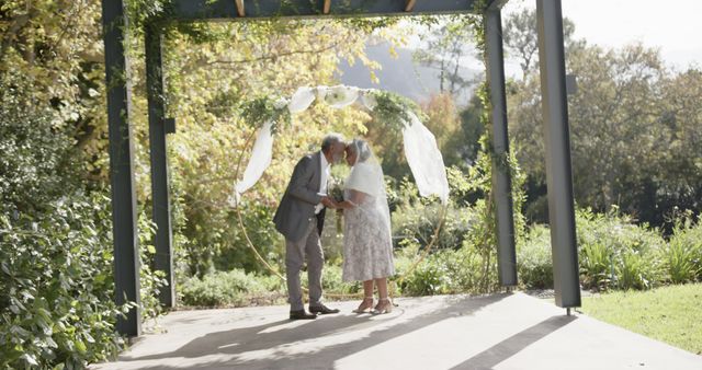 Happy diverse senior bride and groom hold hands touching heads at sunny outdoor wedding ceremony. Marriage, romance, retirement and senior lifestyle, unaltered