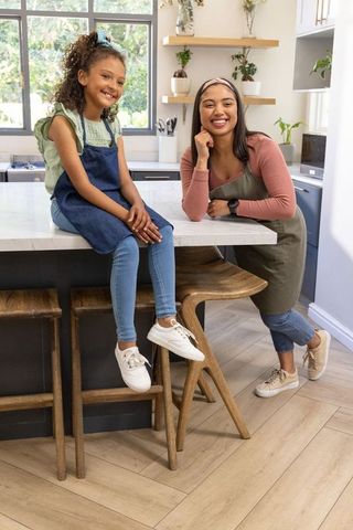Smiling mother and daughter posing on marble kitchen island in denim aprons and sneakers