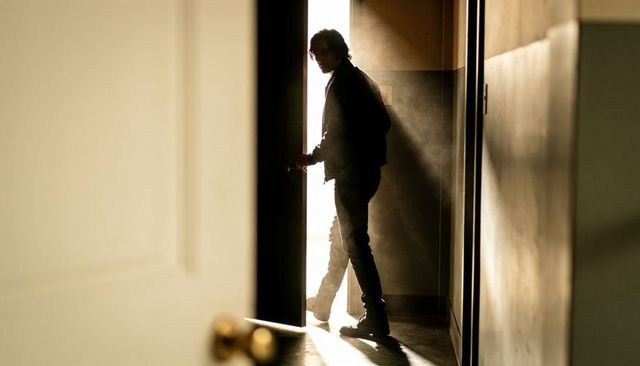 Silhouetted man stepping through backlit doorway touching brass knob with dust beam