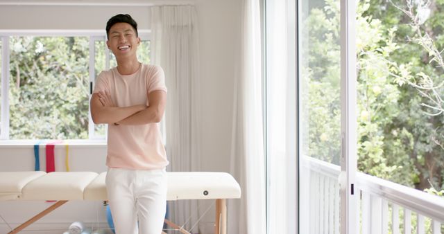 Young man standing indoors in a bright living space with large windows, enjoying natural light. Ideal for concepts related to home leisure, positivity, modern living, interior design, and casual lifestyle promotions.