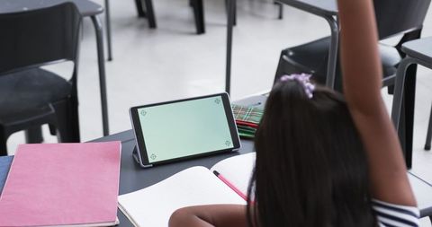 Asian Girl Raising Hand in Classroom Next to Tablet and Books